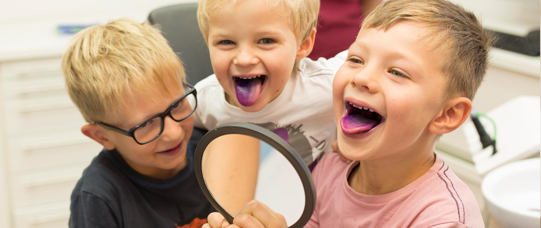 Drei lachende Kinder mit gefärbten Zungen in der Zahnarztpraxis.