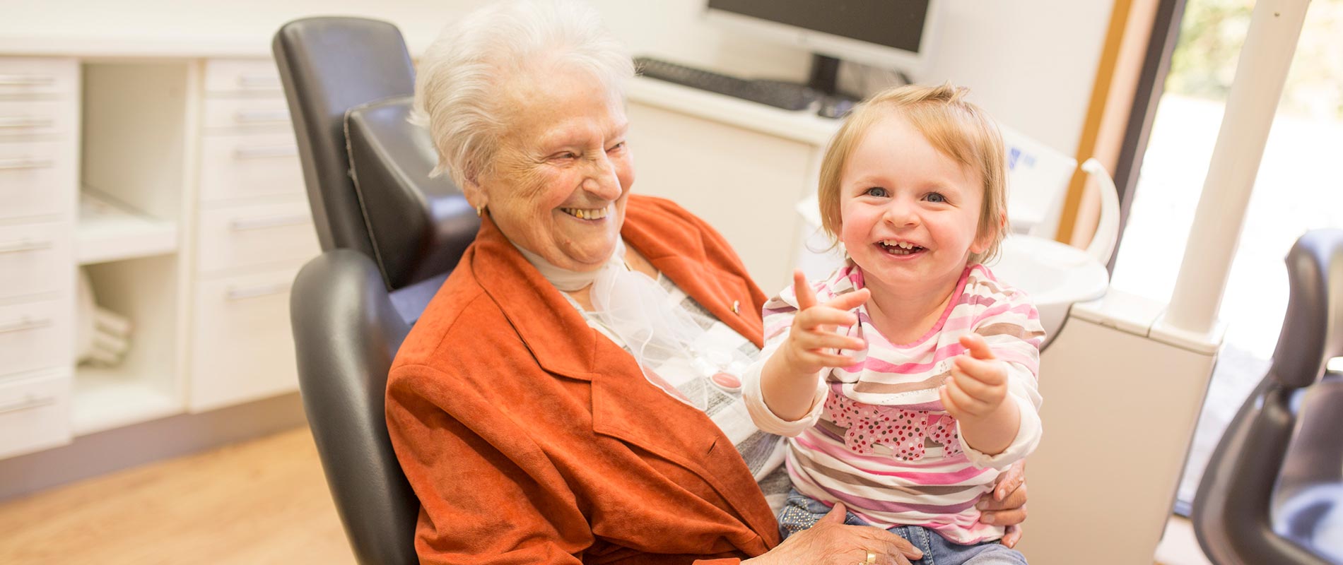 Ältere Patientin mit Enkelkind im Zahnarztstuhl, beide lächeln.