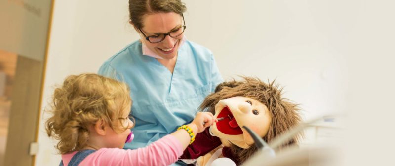 Zahnärztin zeigt Kind mit Puppe richtige Zahnpflege.