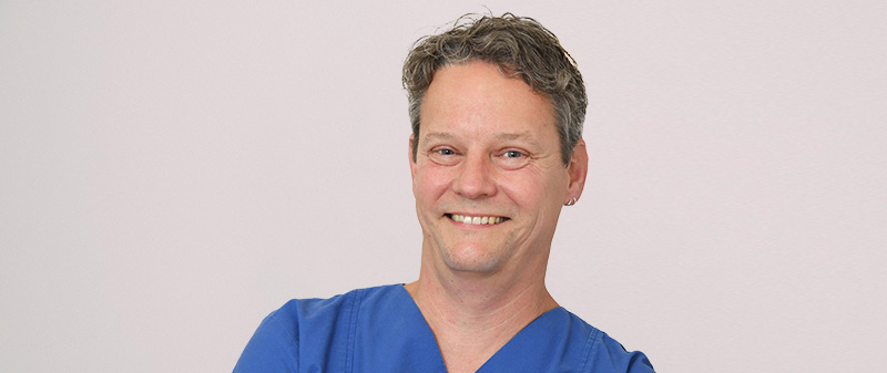 Zahnarzt in blauer Arbeitskleidung lächelt in die Kamera.